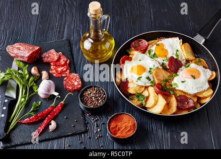 Traditionelle spanische Huevos rotos oder Huevos estrellados - gebratene Eier mit Kartoffeln, Würstchen Chorizo in einer Pfanne auf einem schwarzen Holztisch mit Ingr Stockfoto