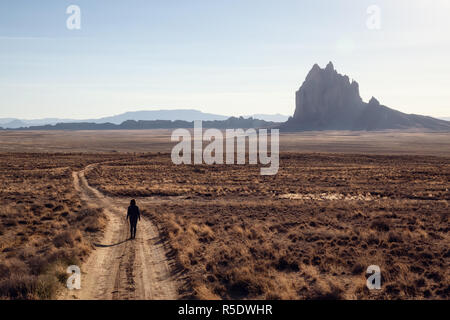 Frau, die zu Fuß auf einem Feldweg in der trockenen Wüste mit einem Berg im Hintergrund. Bei Shiprock, New Jersey, USA. Stockfoto