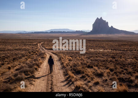 Frau, die zu Fuß auf einem Feldweg in der trockenen Wüste mit einem Berg im Hintergrund. Bei Shiprock, New Jersey, USA. Stockfoto