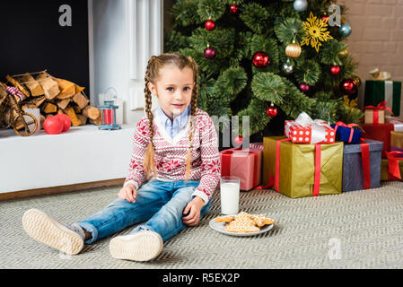 Adorable Kind mit Lebkuchen cookies und Glas Milch sitzen in der Nähe von Weihnachtsbaum und Kamera Stockfoto