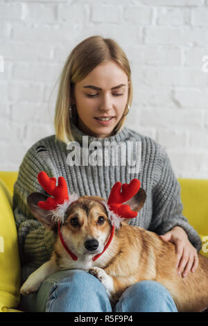 Glückliche junge Frau sitzt auf der Couch und Durchführung corgi Hund mit Weihnachten Hirsch Hörner Stockfoto