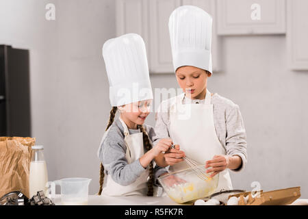 Kleine Kinder in Kochmützen und Schürzen Eier rühren in der Schüssel am Tisch in der Küche Stockfoto