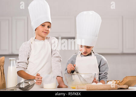 Kleine Jungen an der Kamera schaut und seine Schwester rühren, Eier in der Schale am Tisch in der Küche Stockfoto