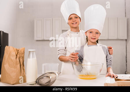 Glückliche Schwester und Bruder in Schürzen und kochmützen Eier rühren in der Schüssel am Tisch in der Küche Stockfoto