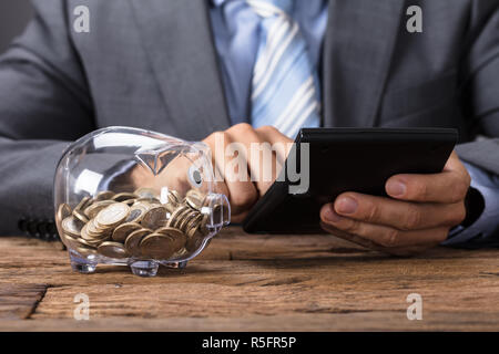 Geschäftsmann mit Taschenrechner durch Glas Sparschwein am Tisch Stockfoto