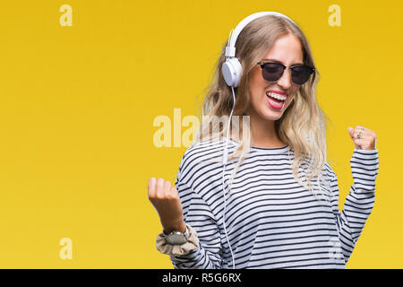 Schöne, junge blonde Frau mit Kopfhörern und Sonnenbrillen über isolierte Hintergrund sehr glücklich und aufgeregt, Sieger Geste mit erhobenen Armen, Stockfoto