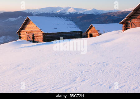 Winter im Bergdorf. Alte Holzhütten auf dem Hügel. Sonnige frostigen Morgen. Blick auf schneebedeckte Berge Stockfoto