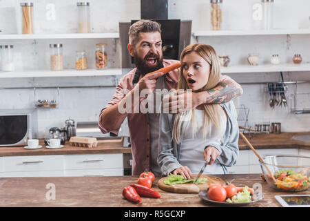 Lustiger Mann, Karotte in der Nähe von Frau Kopf, während Angst Mädchen kochen Abendessen Stockfoto