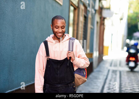 Junge schwarze Mann legere Kleidung Lächeln zu Fuß die Straße runter. Tausendjährigen afrikanischen Kerl mit Latzhose im Freien Stockfoto