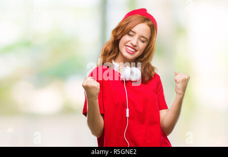 Junge schöne hipster Frau über isolierte Hintergrund Kopfhörer tragen und Kappe sehr glücklich und aufgeregt, Sieger Geste mit erhobenen Armen, smilin Stockfoto