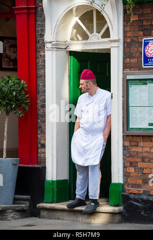 Mann (off-duty Küchenchef in weiß Chef's) stehen vor der Tür außerhalb Restaurant & Hotel, entspannt und die Hände in den Taschen - York, North Yorkshire, England, UK. Stockfoto