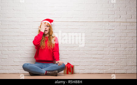 Junge rothaarige Frau über Ziegel Wand saß tragen Mütze Abdecken eines Auges mit der Hand mit selbstbewussten Lächeln auf dem Gesicht und Überraschung Emotion. Stockfoto