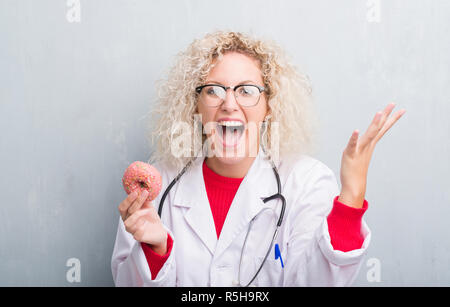 Junge blonde dietist Arzt Frau über Grunge graue Wand holding Donut sehr glücklich und aufgeregt, Sieger Ausdruck feiern Sieg schreien mit großen Stockfoto