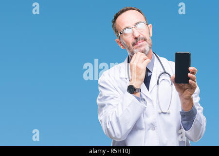 Mittleres Alter senior hoary Arzt Mann zeigt der Bildschirm des Smartphones über isolierte Hintergrund ernstes Gesicht denken über Frage, sehr verworrene Vorstellung Stockfoto