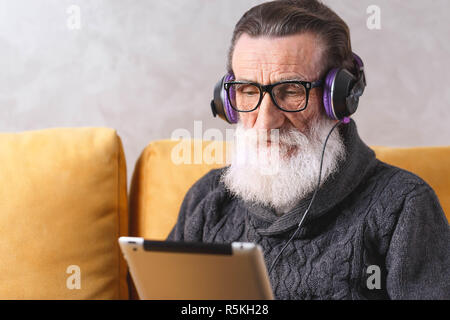 Senior zeitgenössische bärtigen Mann mit Brille grau Pullover sitzen in der Kopfhörer auf ein gelbes Sofa in seinem Lichte Wohnzimmer und interessanten Film auf dem Tablet-PC. Stockfoto