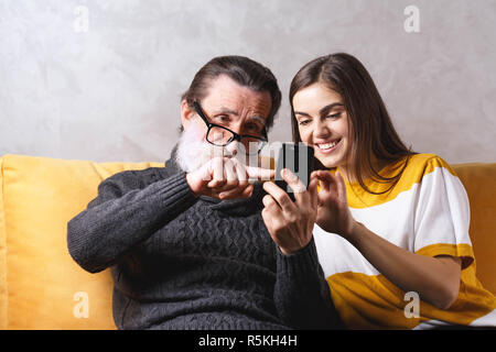 Älteren bärtigen Vater in Gläsern in Anbetracht der Smartphone beim Sitzen auf dem gelben Sofa im hellen Wohnzimmer mit seinem erwachsenen Langhaarige brünette Tochter, moderne Technik, Kommunikation Konzept Stockfoto