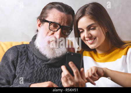 Portrait von älteren bärtigen Vater in Gläsern in Anbetracht der Smartphone beim Sitzen auf dem gelben Sofa im hellen Wohnzimmer mit seinem erwachsenen Langhaarige brünette Tochter, moderne Technik, Kommunikation Konzept Stockfoto