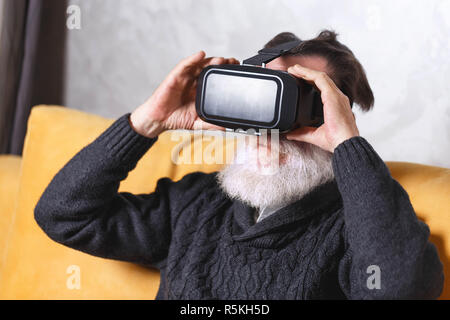 Zeitgenössische älterer bärtiger Mann im grauen pullover Tragen einer Virtual reality Headset beim Sitzen auf dem gelben Sofa im Wohnzimmer, zukünftige Technologie Konzept Stockfoto