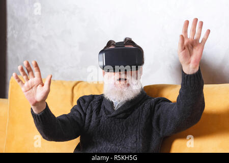 Zeitgenössische älterer bärtiger Mann im grauen pullover Prüfung VR-Gerät während der Sitzung auf dem gelben Sofa im Wohnzimmer, zukünftige Technologie Konzept Stockfoto