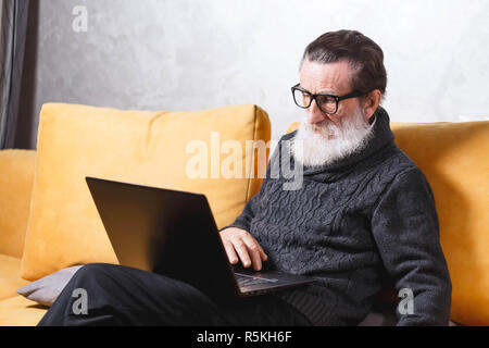 Gut aussehender älterer bärtiger Mann in Gläsern und grauen pullover sitzt mit Laptop auf dem gelben Sofa im Wohnzimmer, moderne Technologie, Kommunikation Konzept Stockfoto