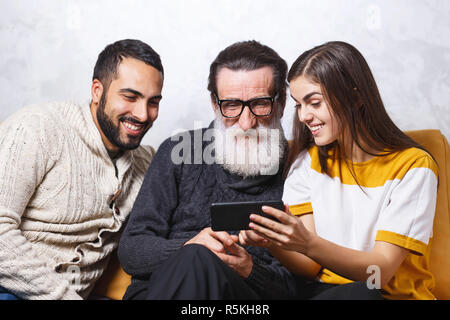 Brunette bärtigen Sohn und Brunette langhaarigen Tochter zeigt Fotos der Ferienhäuser sie älteren bärtigen Vater in Gläser beim Sitzen auf dem gelben Sofa im Wohnzimmer, moderne Technologie, Kommunikation Konzept Stockfoto