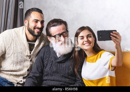 Brunette bärtigen Sohn und Brunette langhaarigen Tochter lächelnd und mit selfie sie älteren bärtigen Vater in Gläser beim Sitzen auf dem gelben Sofa im Wohnzimmer, moderne Technologie, Kommunikation Konzept Stockfoto