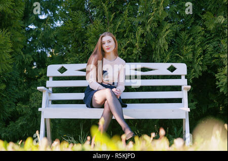 Ein nettes Mädchen mit langen roten Haaren, sitzt auf einer weißen Bank vor dem Hintergrund eines Gartens mit Lebensbäume und lächelt. Porträt im freien Stockfoto