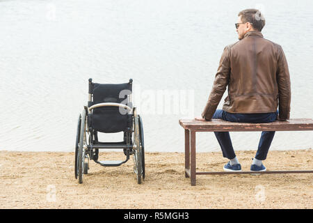 Ein junger Mann sitzt auf einer Bank am See, neben seinem Rollstuhl Stockfoto