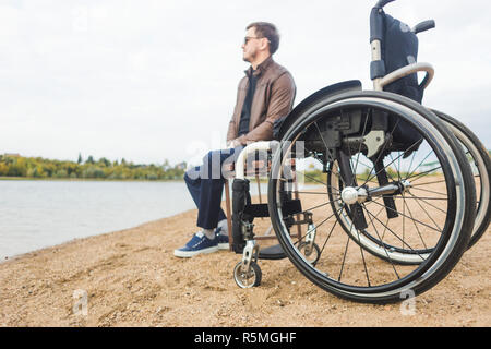 Ein junger Mann sitzt auf einer Bank am See, neben seinem Rollstuhl Stockfoto