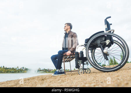 Ein junger Mann sitzt auf einer Bank am See, neben seinem Rollstuhl Stockfoto