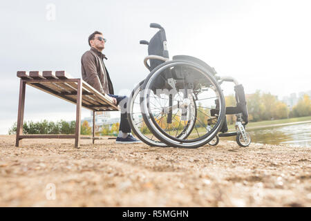 Ein junger Mann sitzt auf einer Bank am See, neben seinem Rollstuhl Stockfoto