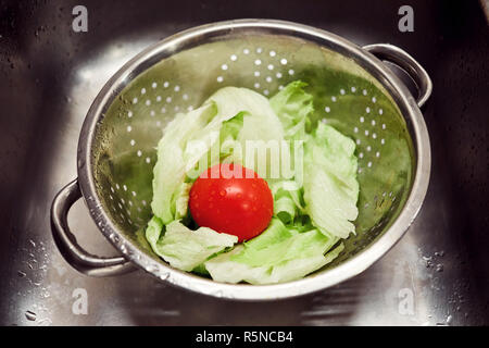 Waschen von frischem Gemüse. Salat und Tomaten in einem silbernen metall Sieb in der Spüle. Stockfoto