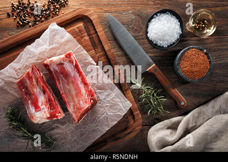 Raw Kalb kurze Rippe auf der weißen kochen Papier und Holz Schneidebrett. Mit Kräutern, Gewürzen und kochmesser eingerichtet. Ansicht von oben. Stockfoto