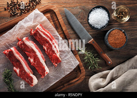 Raw Kalb kurze Rippe auf der weißen kochen Papier und Holz Schneidebrett. Mit Kräutern, Gewürzen und kochmesser eingerichtet. Ansicht von oben. Stockfoto