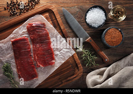 Raw Kalb Flanke Rindfleisch auf weißen kochen Papier und Holz Schneidebrett. Mit Kräutern, Gewürzen und kochmesser eingerichtet. Ansicht von oben. Stockfoto