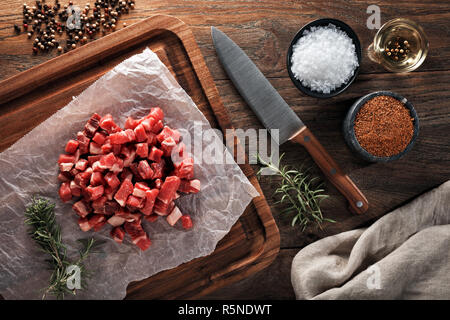 Raw Kalb Fleisch in kleine Stücke auf weißem kochen Papier und Holz Schneidebrett gehackt. Mit Kräutern, Gewürzen und kochmesser eingerichtet. Ansicht von oben. Stockfoto