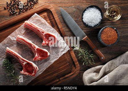 Raw Lamm Rippe Koteletts auf weißem kochen Papier und Holz Schneidebrett. Mit Kräutern, Gewürzen und kochmesser eingerichtet. Ansicht von oben. Stockfoto