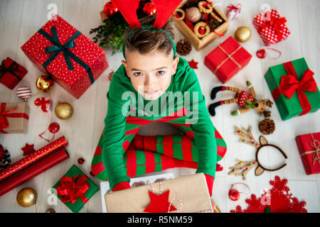 Blick von oben auf eine neugierige Junge tragen xmas Schlafanzug sitzen auf dem Boden, und halte ein Weihnachtsgeschenk, suchen. Stockfoto