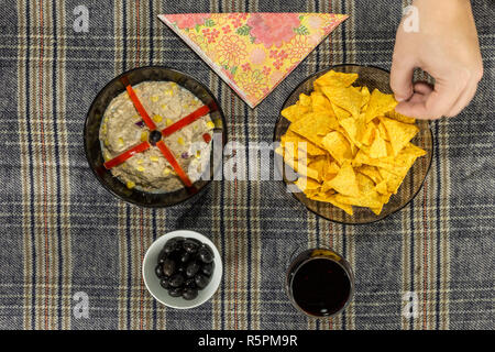 Oben Blick auf eine Schüssel mit Thunfisch Salat, Tortilla Chips, Oliven und Glas Rotwein. Hand nehmen einen Chip. Stockfoto