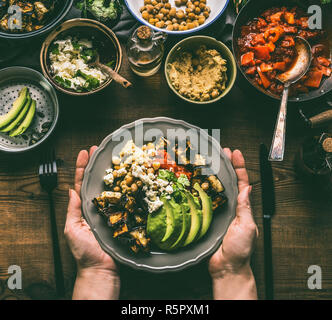 Weibliche Hände halten mit gesunden vegetarischen Schüssel mit verschiedenen gegrilltes Gemüse, Avocado und Kichererbsen Hummus auf rustikalen Hintergrund, Ansicht von oben. Reinigen Stockfoto