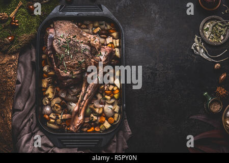 Kochen Vorbereitung der hirschbraten. Bein der Hirsch mit Knochen in gusseisernen Pfanne mit Darm Gemüse auf dunklen Küche Tisch Hintergrund mit Kräutern und Gewürzen Stockfoto