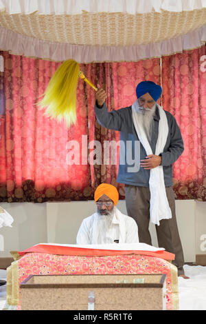 Ein Sikh Priester liest aus der Heiligen Gebetbuch, während eine andere devotee Wellen eine gelbe Chaur Sahib an einem Tempel in South Richmond Hill, Queens, New York. Stockfoto