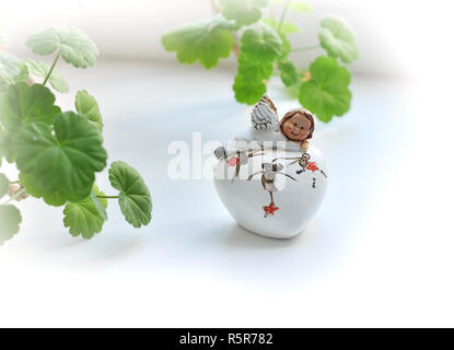 Baby Engel Statuette bleiben auf dem Herzen. Weihnachten Engel in der Nähe der Fenster. Pelargonium Blumen Topf auf dem Fenster und einer Figur aus einem niedlichen Engel Stockfoto