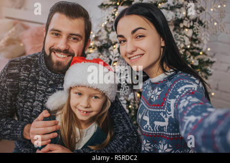 Lächelnde junge Familie in Weihnachtsstimmung, Foto mit Smartphone. Stockfoto