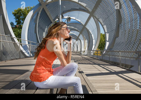 Frau mit orangefarbenen T-Shirt im Sommer, bei modernen städtischen Steg sitzen, telefonieren mobil, in Madrid, Spanien, Europa Stockfoto