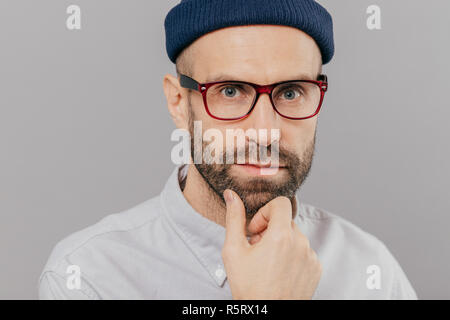 Nahaufnahme von Stattlichen bärtiger junger Mann hält Kinn, sieht Ernst direkt an der Kamera, denkt an etwas, trägt eine Brille, weiße Kleidung, ich Stockfoto