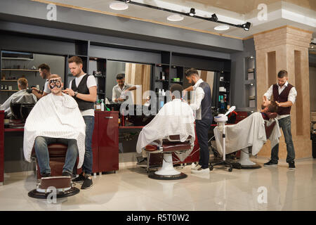 Ansicht von der Rückseite der drei Friseure im weißen T-Shirts und Westen Wartung männlichen Kunden in den Friseur. Professionelle Friseure Haare schneiden, den Bart rasieren und Gesichtsmaske. Stockfoto