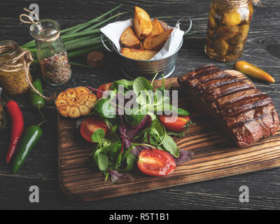 Gegrilltes Steak auf dem Brett serviert mit Kräutern, Bratkartoffeln, Gewürze auf einem schwarzen Oberfläche Stockfoto