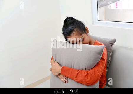 Traurig asiatische Frau umarmen, ein Kissen und sitzt auf einem Sofa Stockfoto