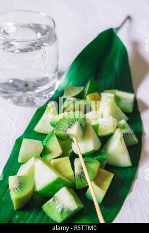 Grüne Früchte mix Salat auf einem großen Blatt und einem Glas Wasser, auf einem weißen Tisch. Detox essen. Vegetarische Gerichte Desserts. Gesunde Ernährung Kontext. Stockfoto